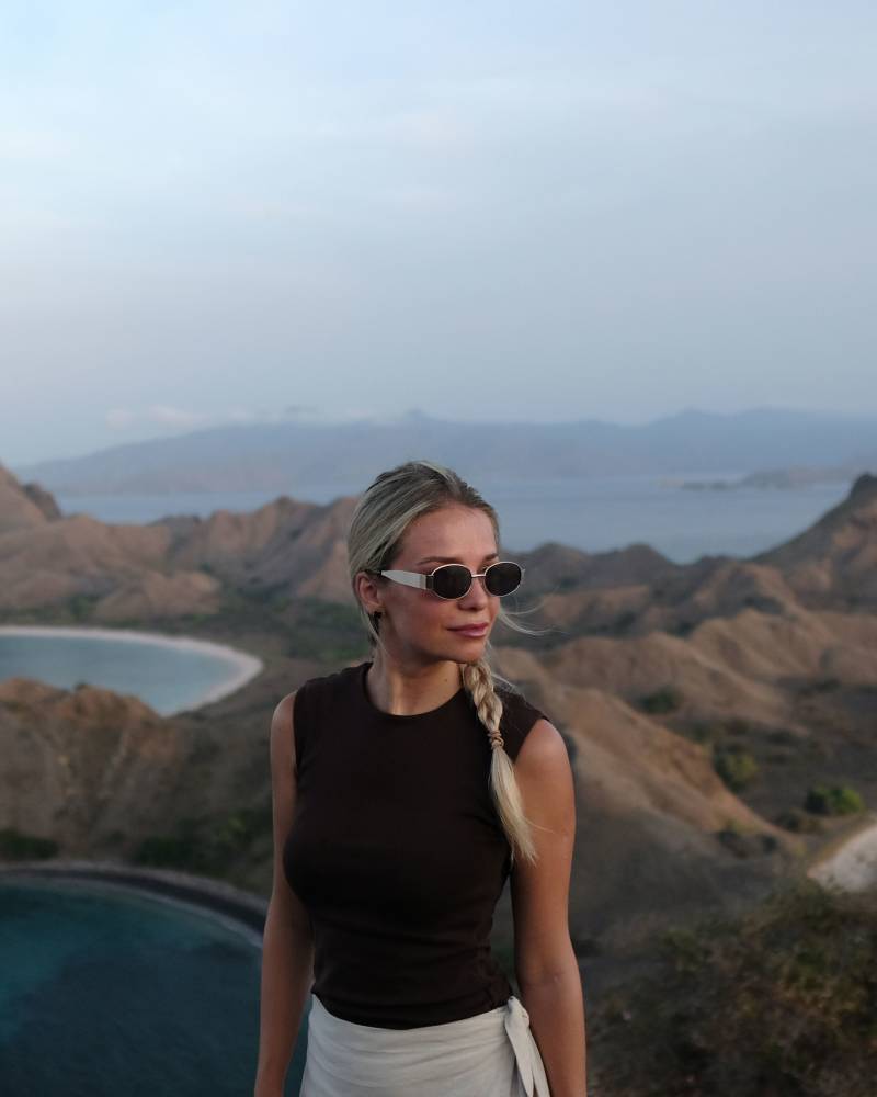 A woman in sunglasses and a brown top stands in the foreground, with a scenic background of rolling hills and calm ocean waters under a hazy sky.