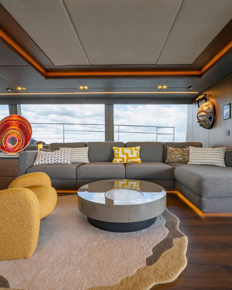 A modern living area with plush yellow armchairs and a sectional sofa in the foreground, and large windows offering a view of the sky and water in the background.