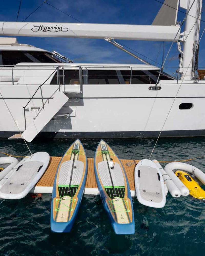 Four paddleboards and two yellow jet skis are docked on a wooden platform in the foreground, with a large white yacht docked behind them.