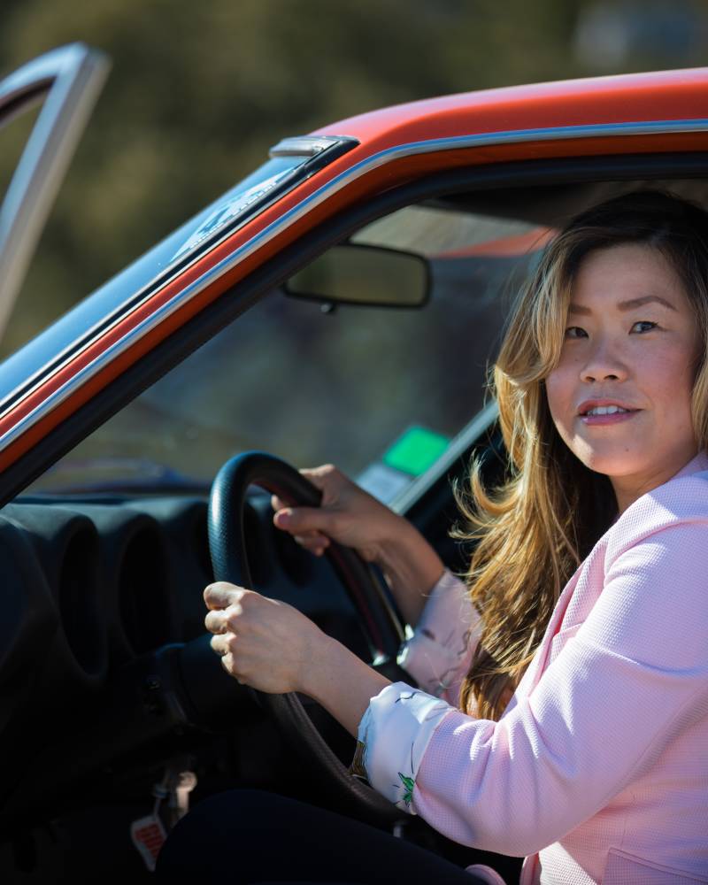 A woman in a pink blazer sits in the driver's seat of an orange car, with a blurred outdoor landscape in the background.
