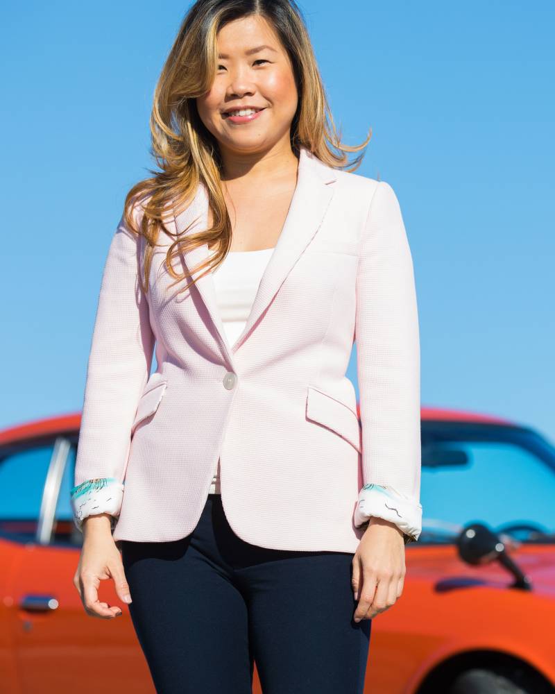 A smiling woman in a pink blazer stands in front of a bright orange car against a clear blue sky.