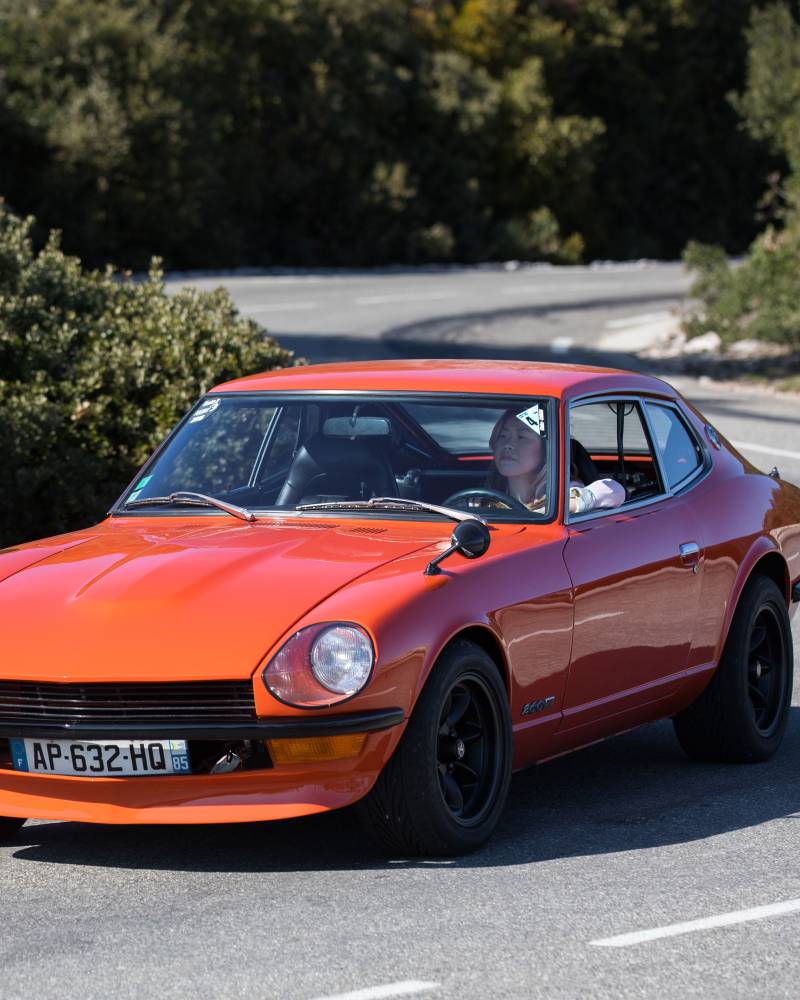 An orange vintage sports car with black rims drives on a paved road, with lush green trees and a clear blue sky in the background.
