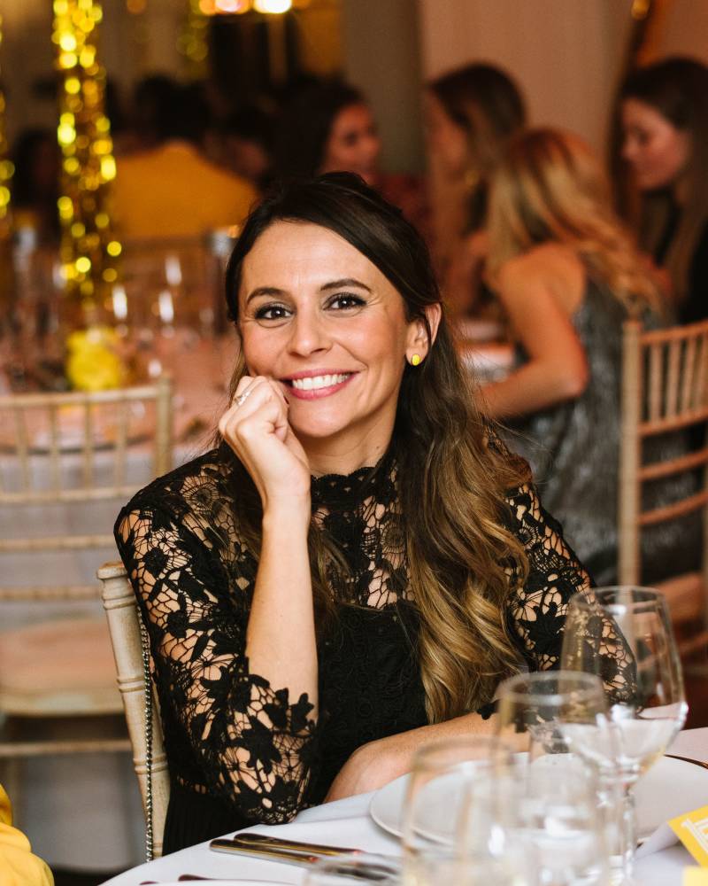 A smiling woman in a black lace dress rests her chin on her hand in the foreground, while other guests and golden decorations are blurred in the background.