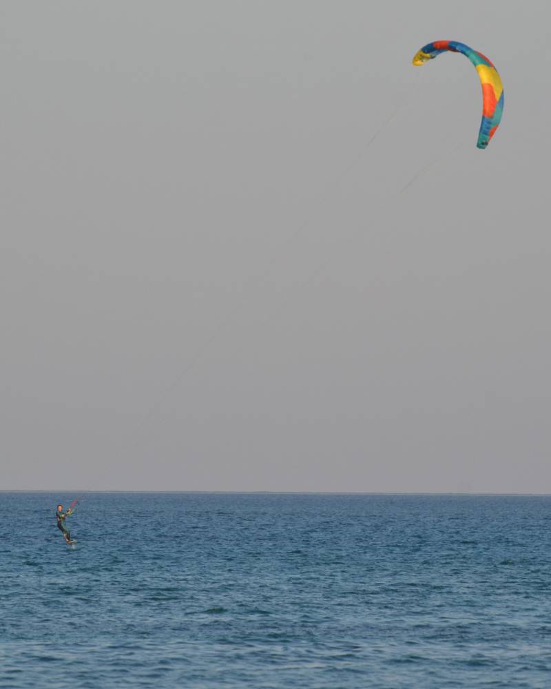 A kite surfer glides across the blue ocean under a pale, overcast sky with a colorful kite soaring high above.