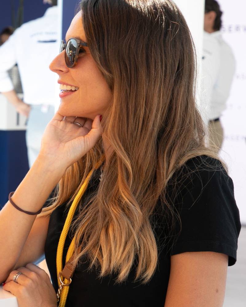 A woman with long brown hair and sunglasses smiles in the foreground, while blurred figures in white uniforms stand in the background.