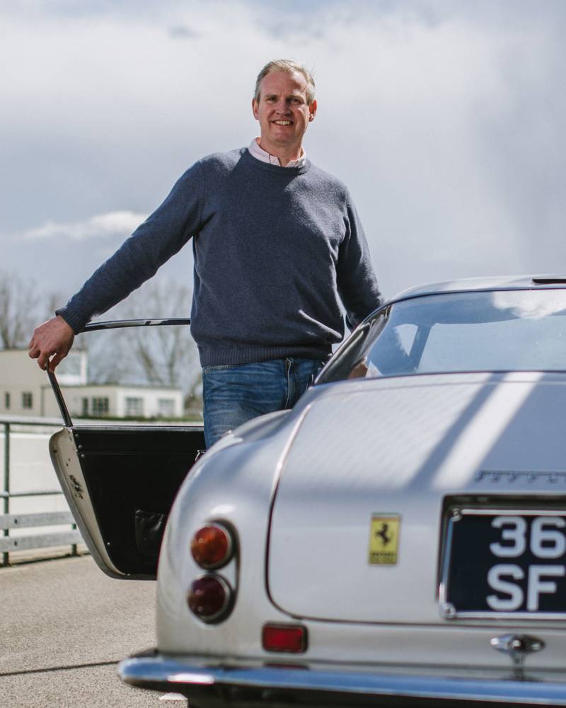 A smiling man in a blue sweater stands beside a silver vintage Ferrari against a cloudy sky and a distant building.