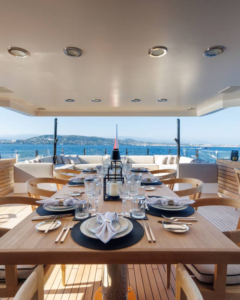 A set dining table with wooden chairs sits on the deck of a luxury yacht, overlooking a calm blue sea and distant coastline.
