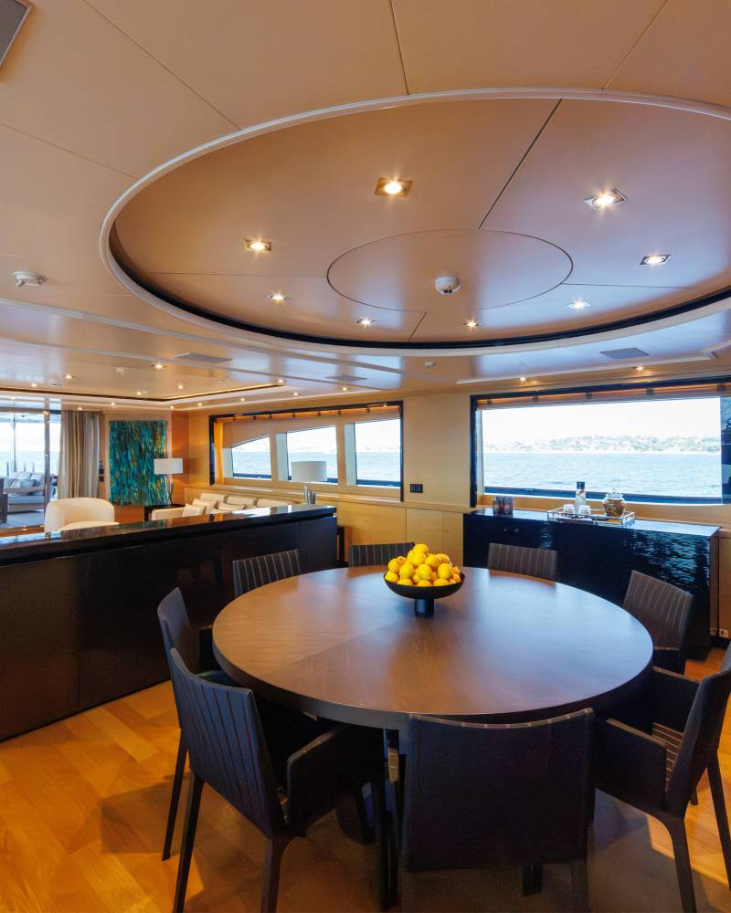 A round wooden dining table with a bowl of lemons sits in the foreground of a luxurious yacht interior, with a lounge area and ocean views visible in the background.