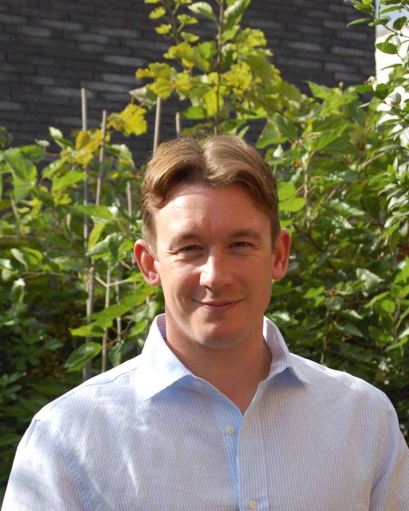 A man in a light blue striped shirt stands in the foreground against a background of lush green foliage and a dark grey wall.