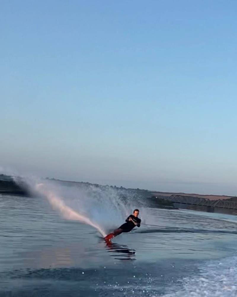 A person wakeboards across a calm body of water, creating a large white spray in the foreground against a backdrop of distant hills and a clear blue sky.