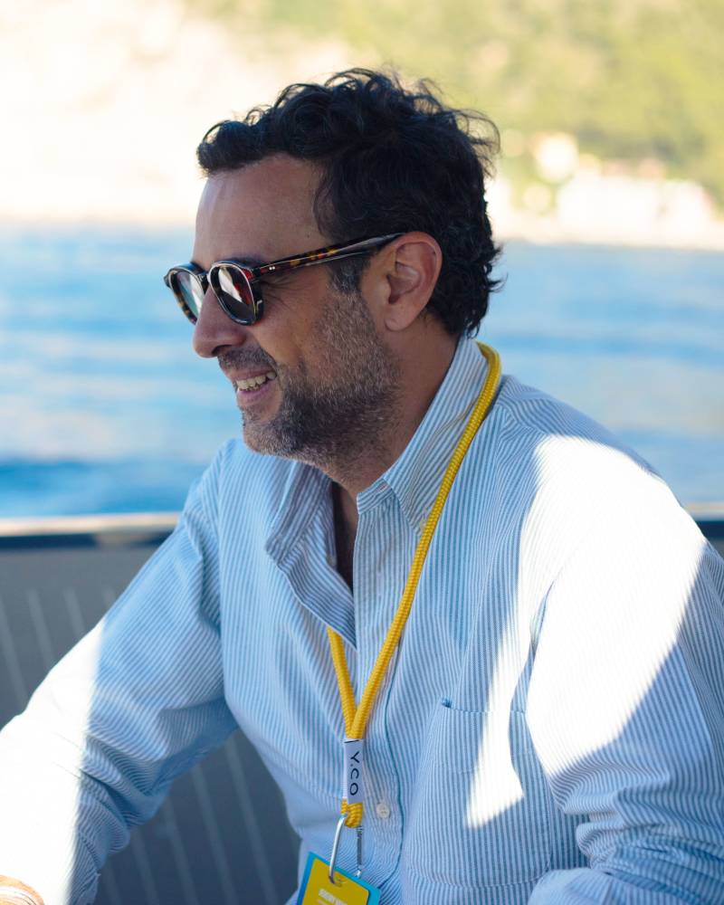 A man wearing sunglasses and a striped blue shirt stands in the foreground against a blurred background of blue water and a sunlit coastline.