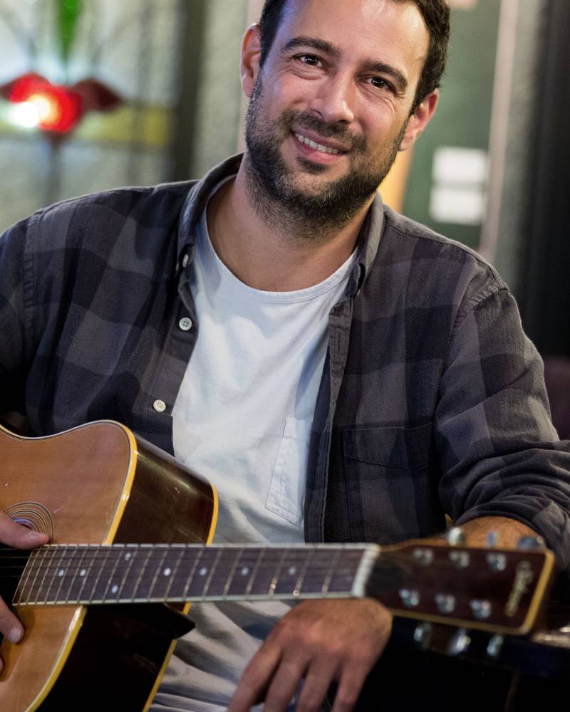 A smiling man in a plaid shirt holds an acoustic guitar in the foreground against a blurred indoor background.