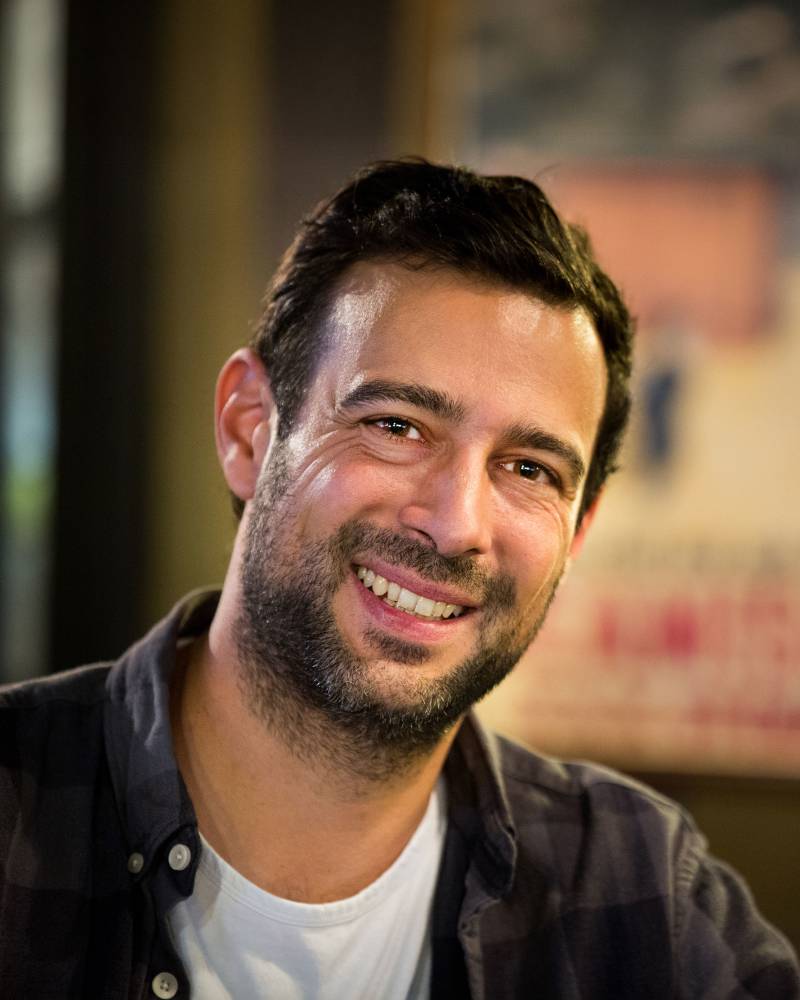 A smiling man with a beard wearing a dark button-down shirt over a white t-shirt stands before a blurred, warm-toned background.