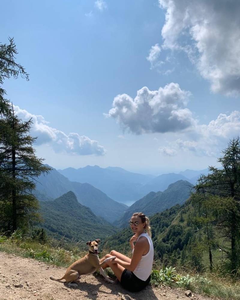A woman and her dog sit on a dirt path in the foreground, overlooking a vast landscape of rolling blue mountains under a cloudy sky.