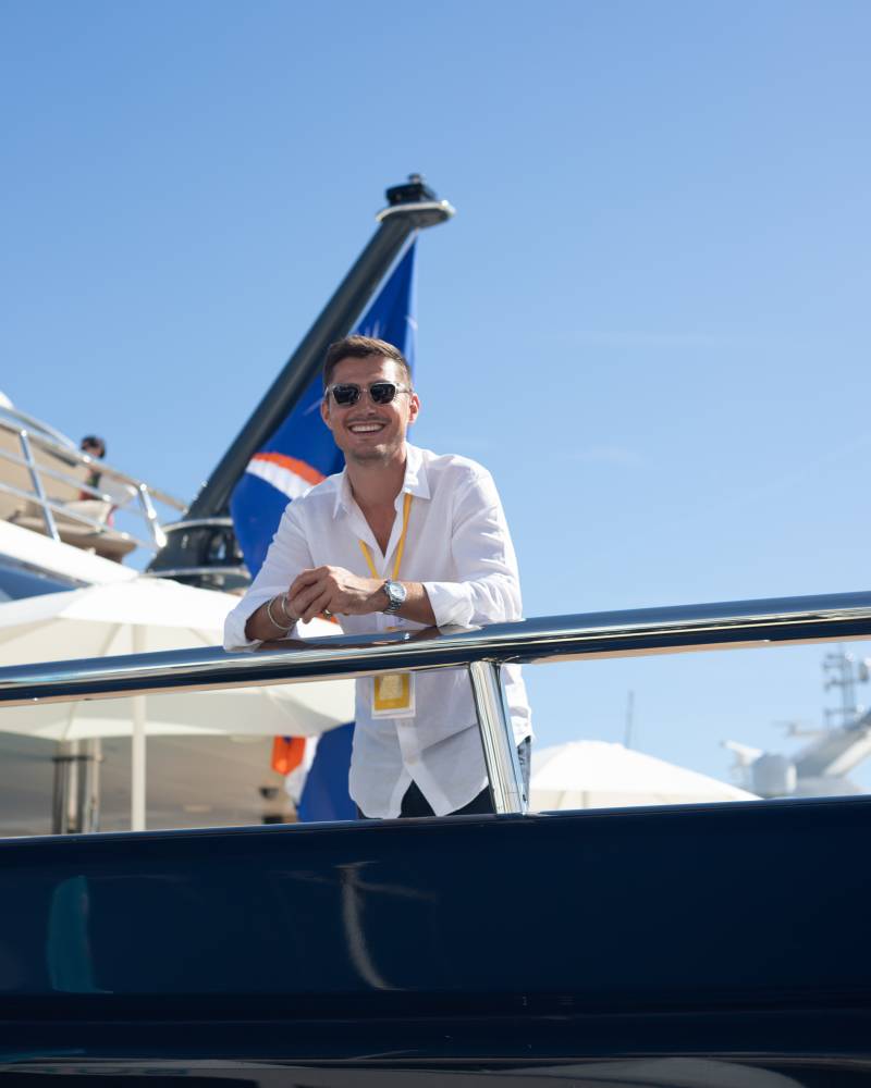 A smiling man in a white shirt and sunglasses leans on a yacht railing against a clear blue sky and a blue flag.