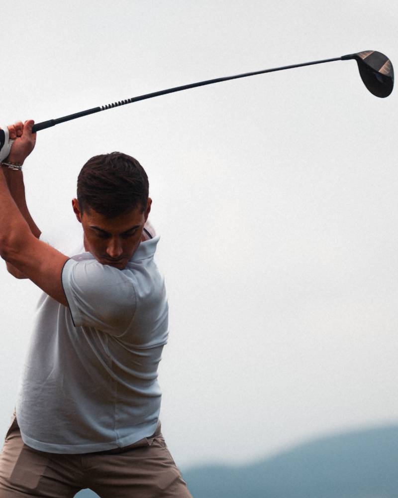 A man in a white shirt swings a golf club against a backdrop of a pale sky and distant mountains.