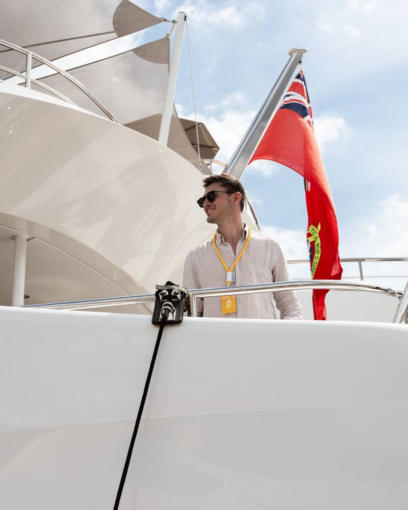 A man in sunglasses and a white shirt stands on a sleek white yacht against a backdrop of a red flag and a cloudy sky.