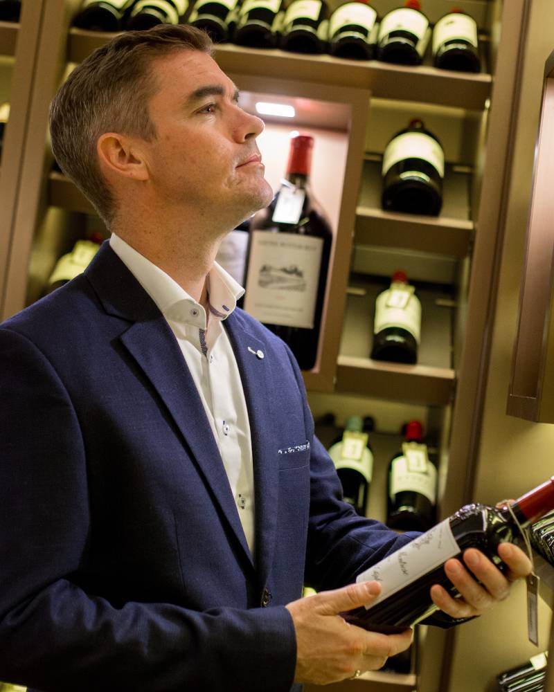 A man in a navy blue suit holds a wine bottle in the foreground, standing before shelves filled with various wine bottles in the background.