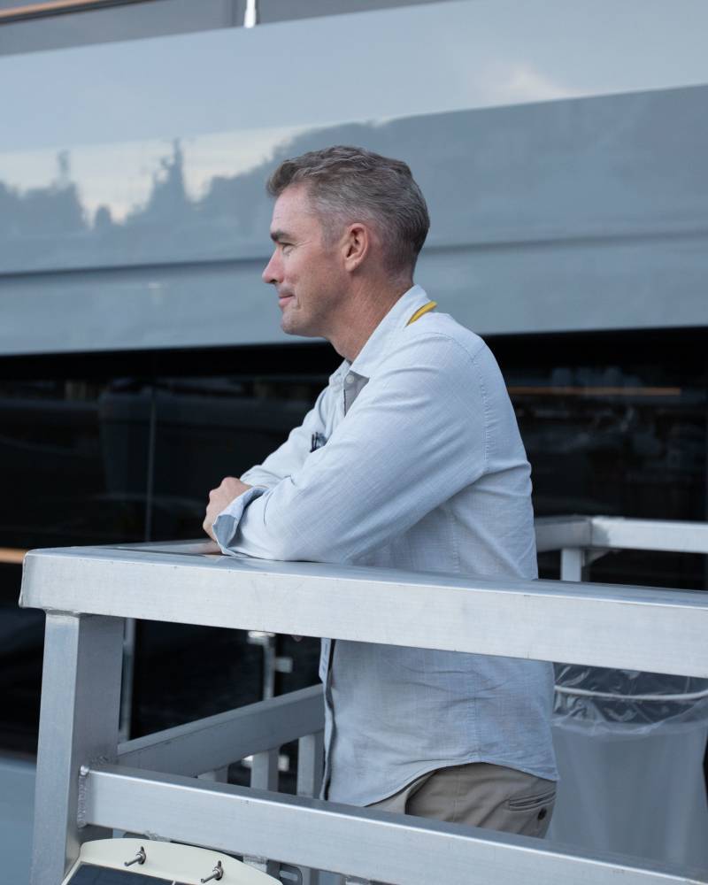 A middle-aged man in a light-colored shirt leans on a silver railing in the foreground, with a large, sleek grey structure and a railing in the background.
