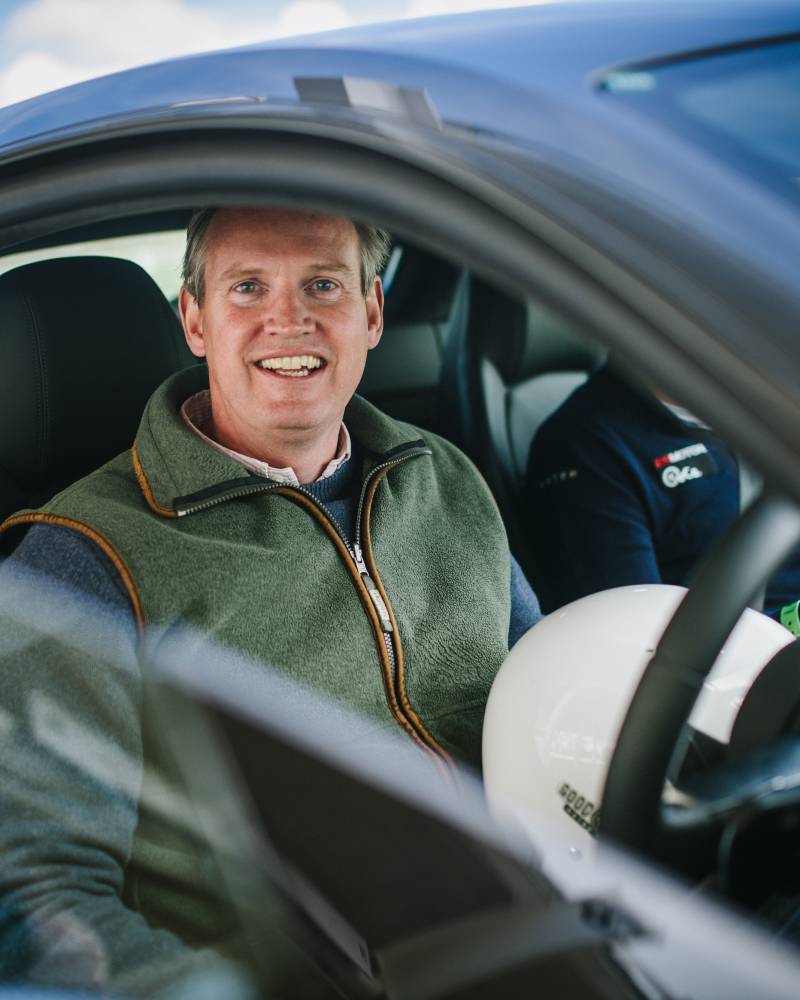 A smiling man in a green vest sits in the driver's seat of a blue car, with another person and a white helmet visible in the background.