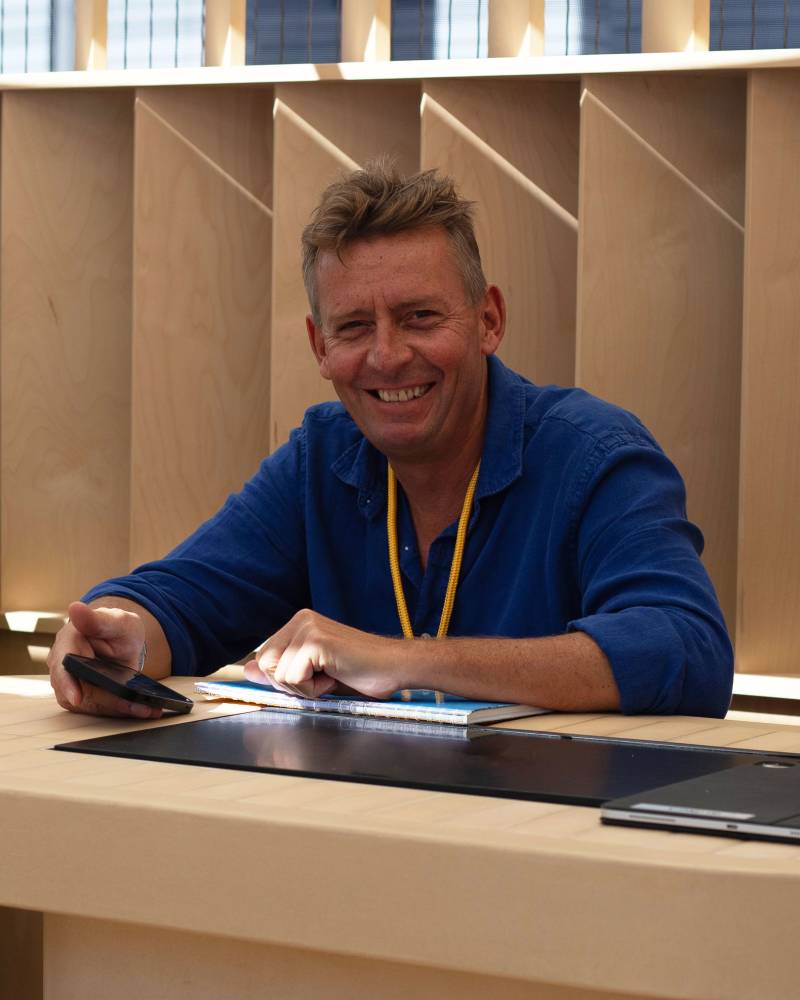 A smiling man in a blue shirt sits at a light-colored wooden desk with a phone and notebook, set against a background of geometric wooden panels.