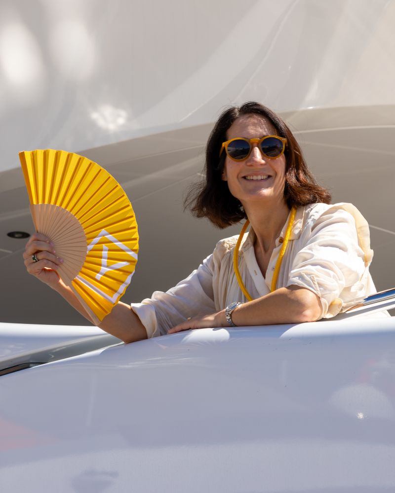 A smiling woman wearing sunglasses and a white shirt holds a yellow fan in the foreground against a sleek, white architectural background.