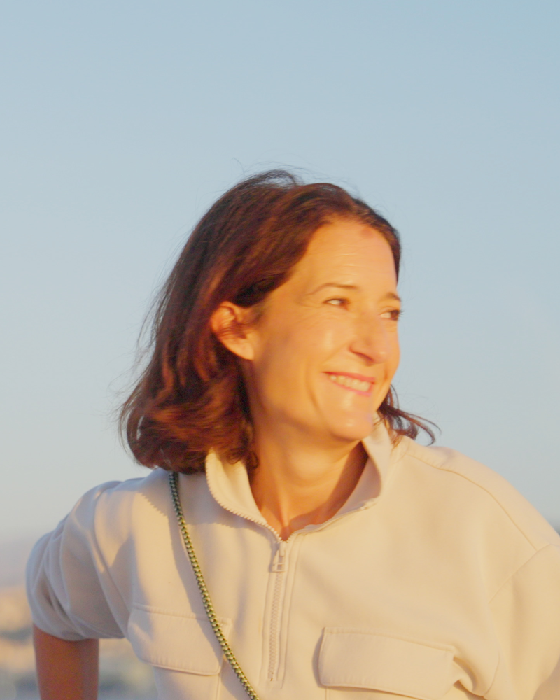 A smiling woman in a beige fleece jacket stands against a clear, pale blue sky.
