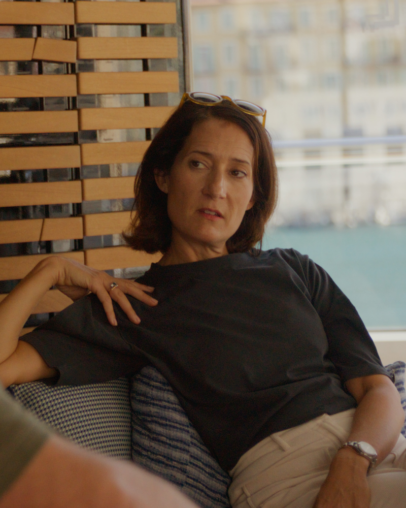 A woman in a dark t-shirt sits on a blue patterned sofa in the foreground, with a wooden slatted wall and a blurred city view in the background.