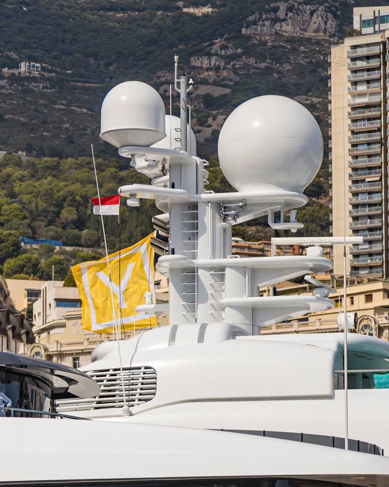 A white luxury yacht with large radar domes stands in the foreground against a backdrop of tall buildings and a steep, forested mountain.