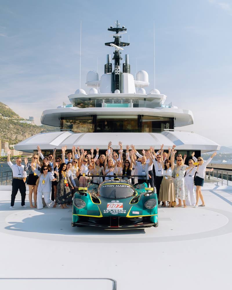 A group of people wave behind a green Aston Martin race car on the deck of a massive white luxury yacht.