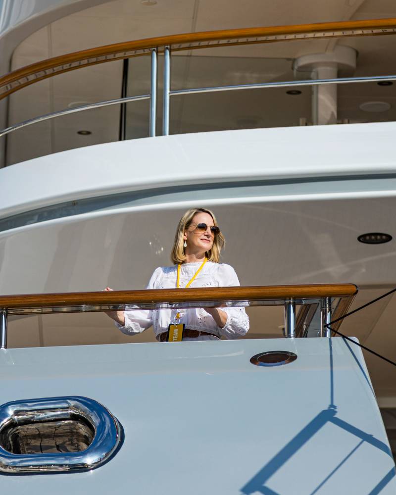 A blonde woman wearing sunglasses and a white blouse stands on the deck of a luxury white yacht.