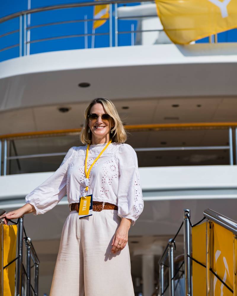 A smiling woman in a white blouse and beige pants stands on a ship's gangway with a white multi-deck vessel and yellow flags in the background.