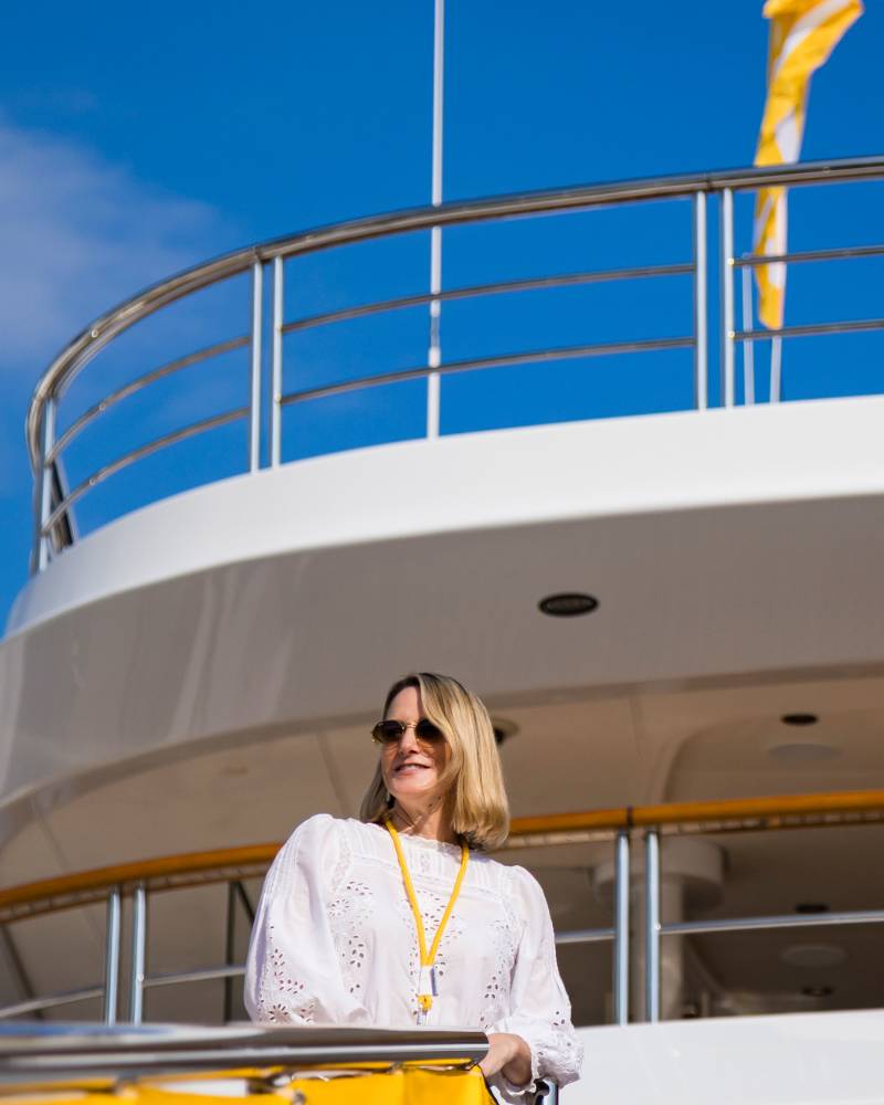 A woman in a white lace blouse and sunglasses stands on a ship's deck against a clear blue sky and white railings.