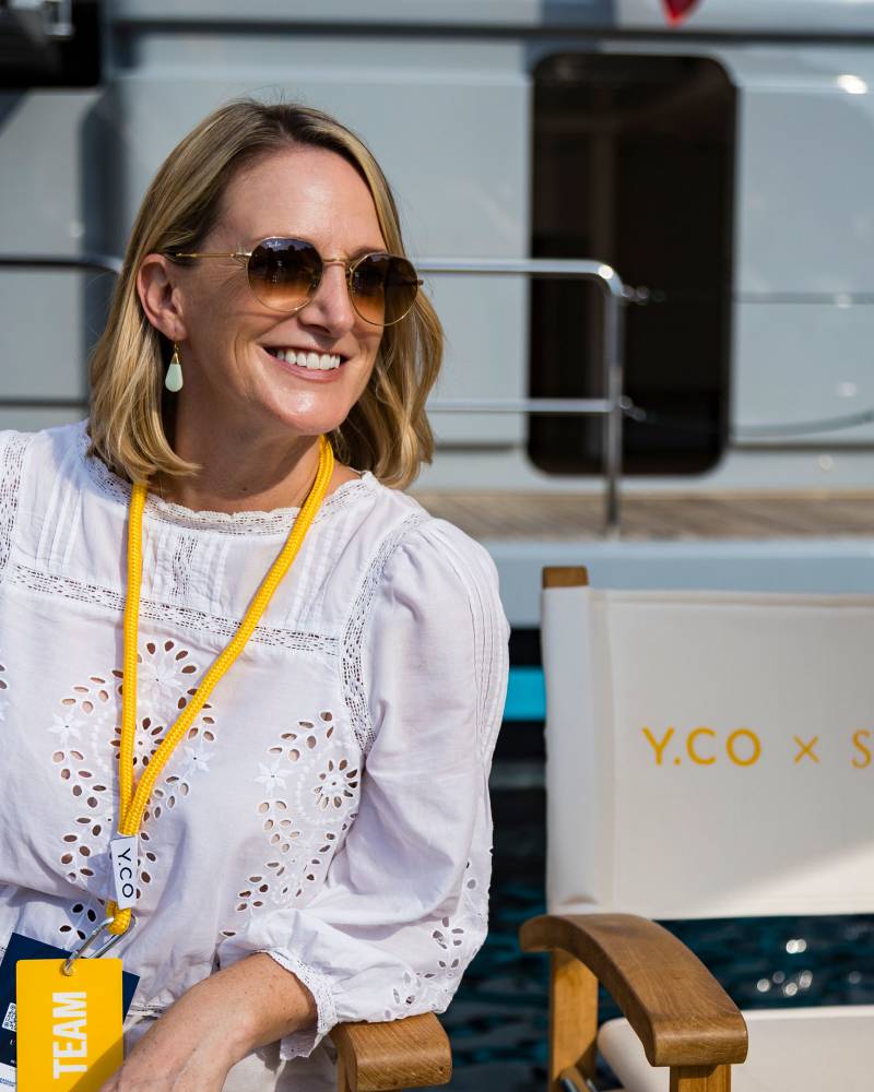 A smiling woman in a white eyelet blouse and sunglasses sits in a deck chair in front of a large white yacht.