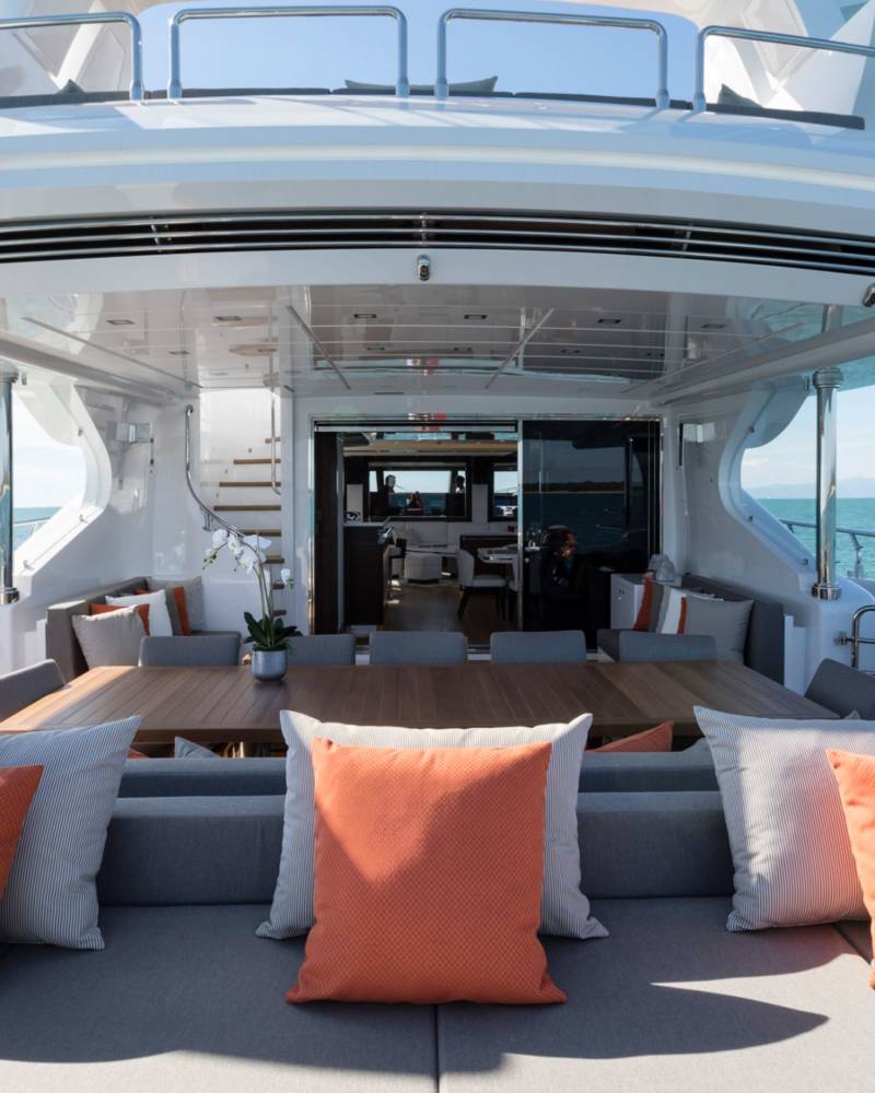 A luxurious yacht deck features a grey lounge sofa with orange pillows in the foreground and a dining area leading to the interior against a backdrop of a clear blue ocean and sky.
