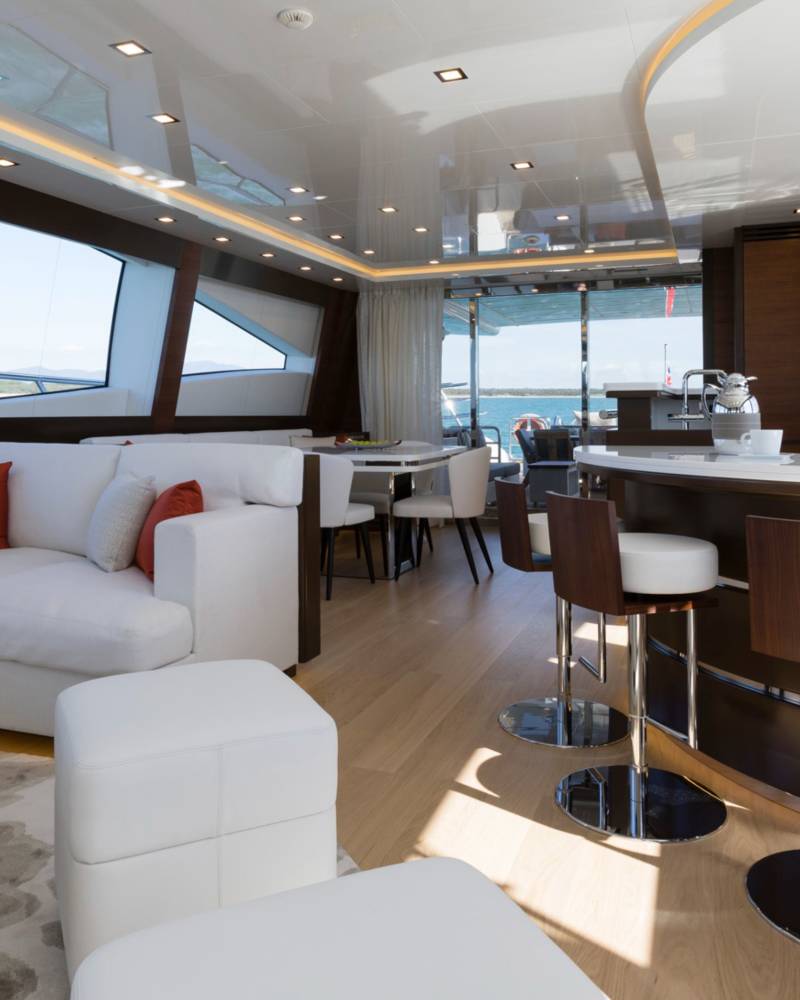 A modern yacht interior features a white sectional sofa and coffee table in the foreground, with a sleek breakfast bar and dining area set against large windows in the background.