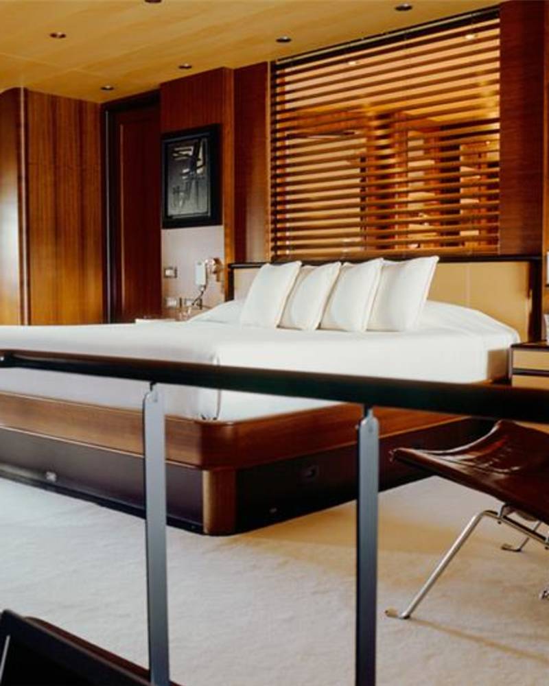 A brown leather chair and black railing sit in the foreground of a luxurious wood-paneled bedroom featuring a large white bed and slatted window blinds.