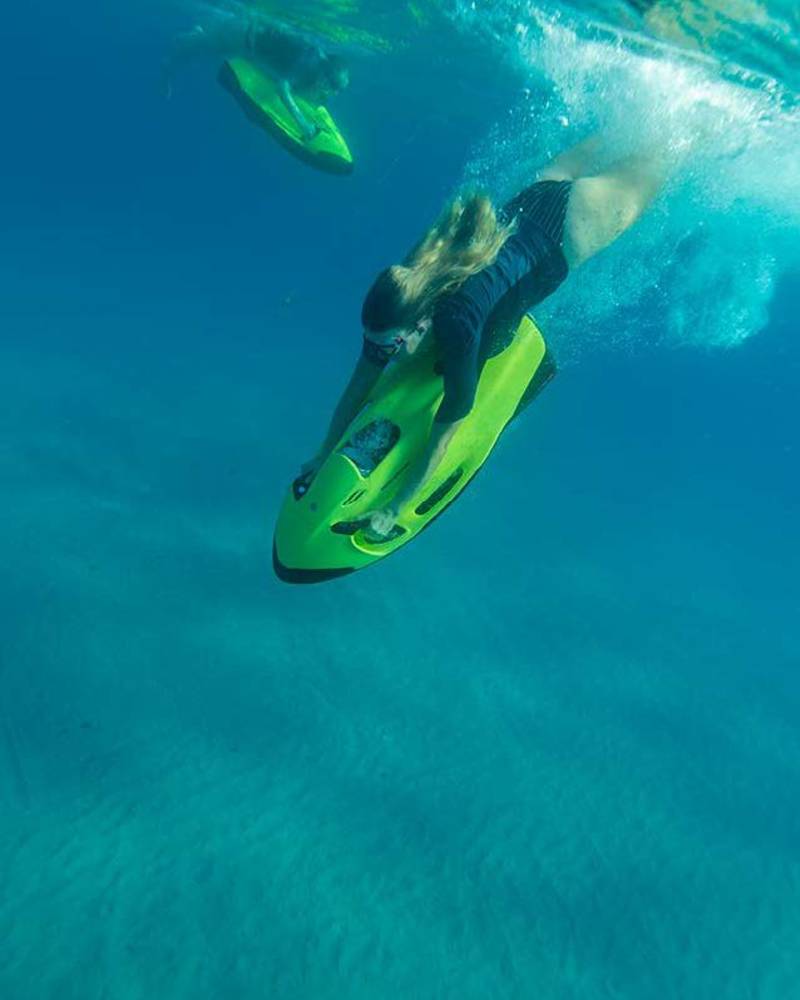 A woman dives underwater on a bright green seabob in the foreground, with another person on a similar device gliding through the blue water in the background.