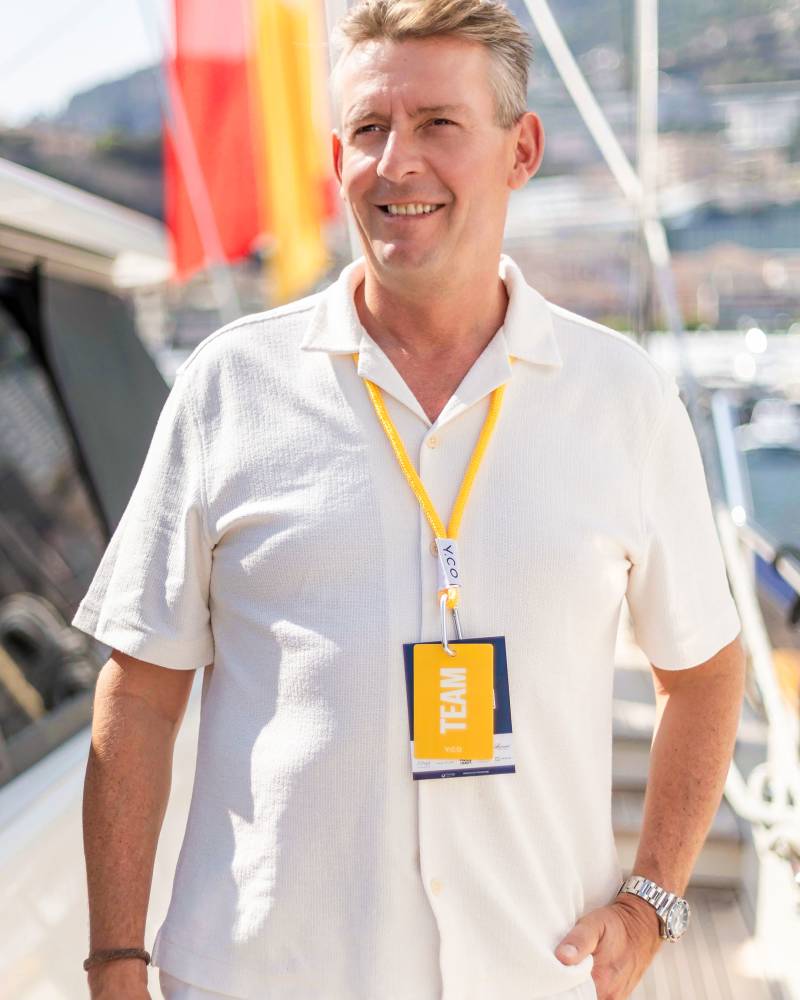A smiling man in a white short-sleeved shirt and lanyard stands in the foreground against a blurred background of a boat and a red and yellow flag.