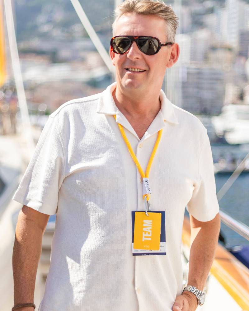 A man wearing sunglasses and a white shirt with a "TEAM" badge stands in the foreground against a blurred background of a harbor and coastal city.