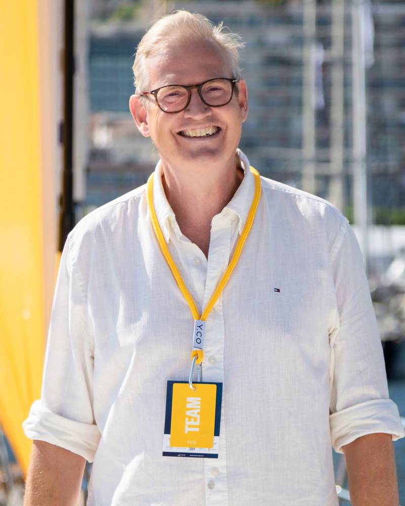 A smiling man wearing glasses and a white shirt with a "TEAM" lanyard stands in the foreground against a blurred background of a harbor and city.