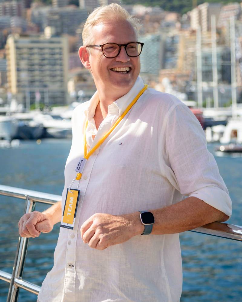 A smiling man in a white linen shirt and glasses leans on a boat railing with a harbor and city skyline in the background.