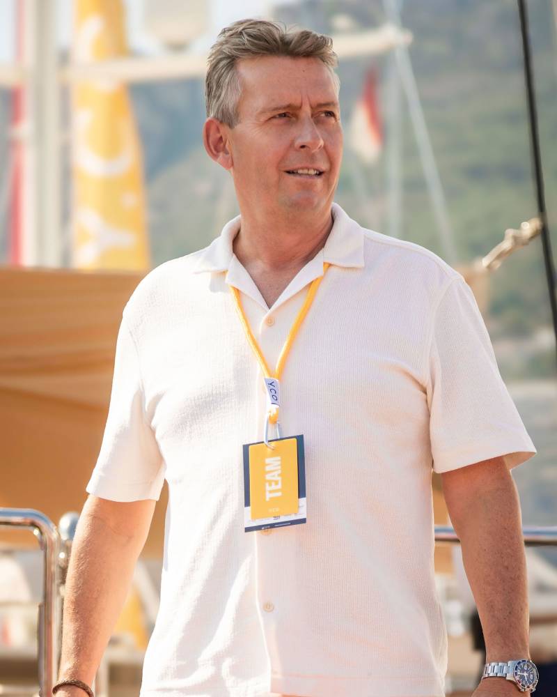 A man wearing a white short-sleeved shirt and a "TEAM" lanyard stands in the foreground against a blurred background of a boat and distant hills.