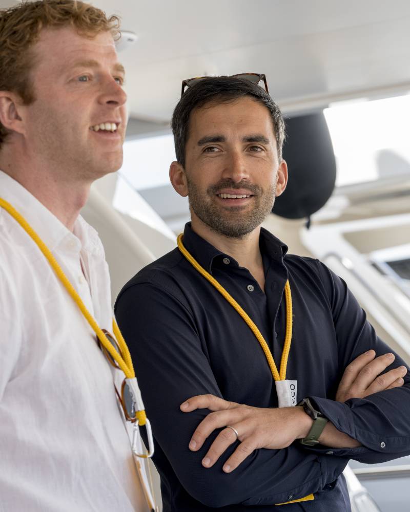 Two men wearing yellow lanyards stand in the foreground against the bright, modern interior of a luxury yacht.