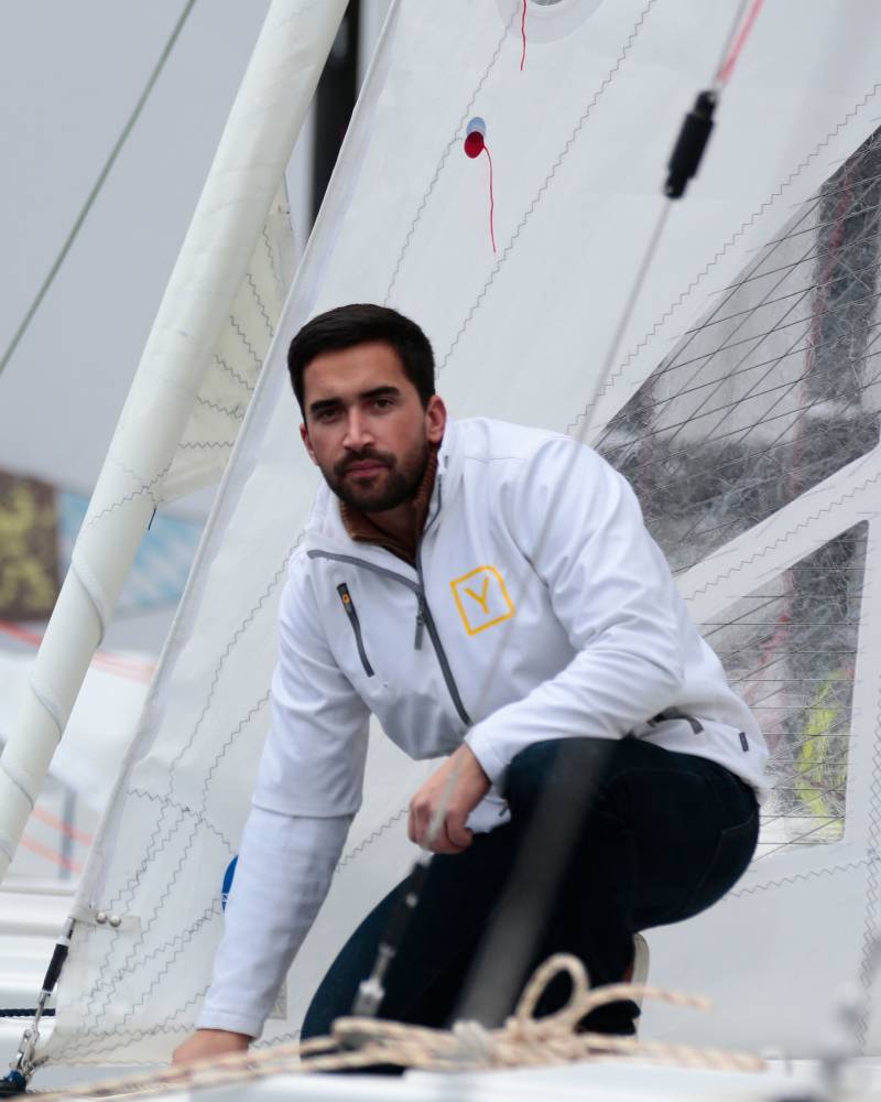 A man in a white jacket and dark pants crouches in the foreground against the backdrop of a white sailboat sail.