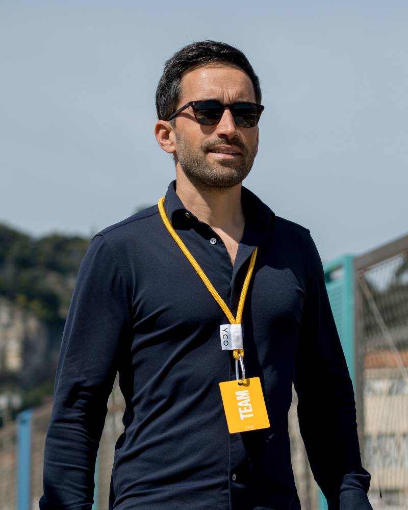 A man wearing sunglasses and a navy blue shirt with a yellow "TEAM" lanyard stands against a blurred background of a hillside and a clear blue sky.