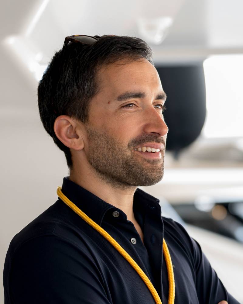 A smiling man with a yellow lanyard stands in the foreground against a blurred, bright white architectural background.