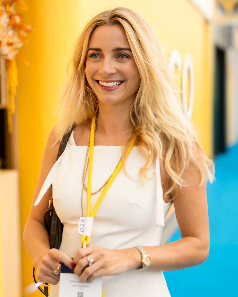 A smiling blonde woman in a white dress and lanyard stands in the foreground against a blurred yellow and blue background.