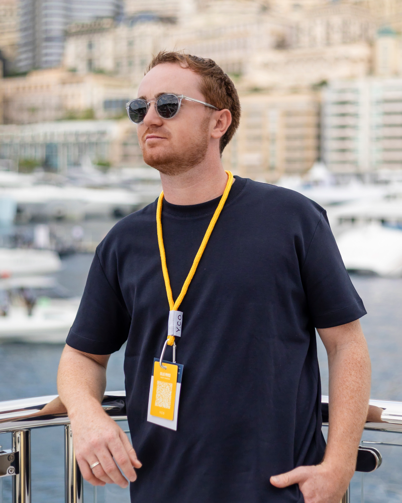 A man wearing sunglasses and a navy blue t-shirt stands by a railing with a blurred cityscape and harbor in the background.