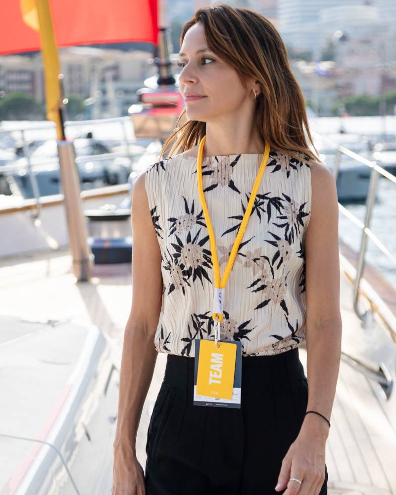 A woman wearing a floral sleeveless top and a "TEAM" lanyard stands on a boat deck with a blurred harbor and city skyline in the background.